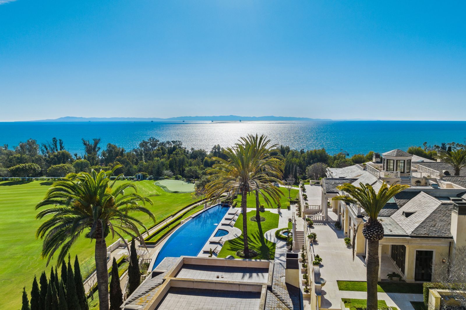 A portion of a Mediterranean mansion, its expansive pool and lawn, and palm trees in the foreground, with the sparkling blue ocean and blue sky on the horizon in the background